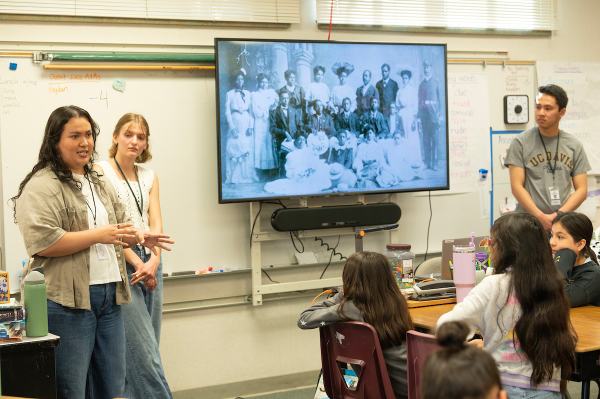 Three students presenting in classroom to seated classmates, large screen behind (photo)