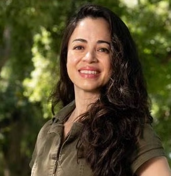 A person with long dark hair, wearing an olive green shirt, smiles in front of lush greenery.