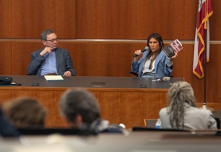 Woman speaking into mic at front of wood-paneled room, man beside her, audience in foreground