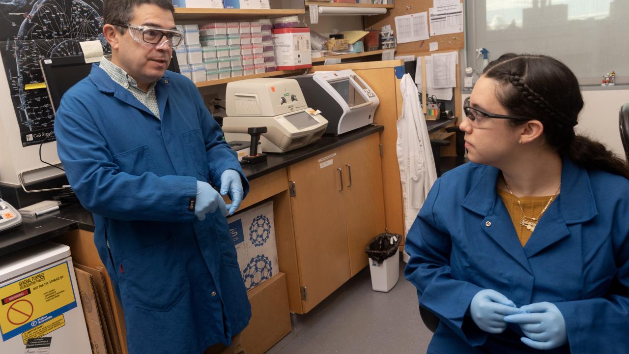 Luis Carvajal-Carmona, Ph.D., associate vice chancellor of academic diversity within the Division of Diversity, Equity and Inclusion, stands in his lab talking to one of his student lab assistants who is seated. Both are wearing a lab coat and lab goggles.