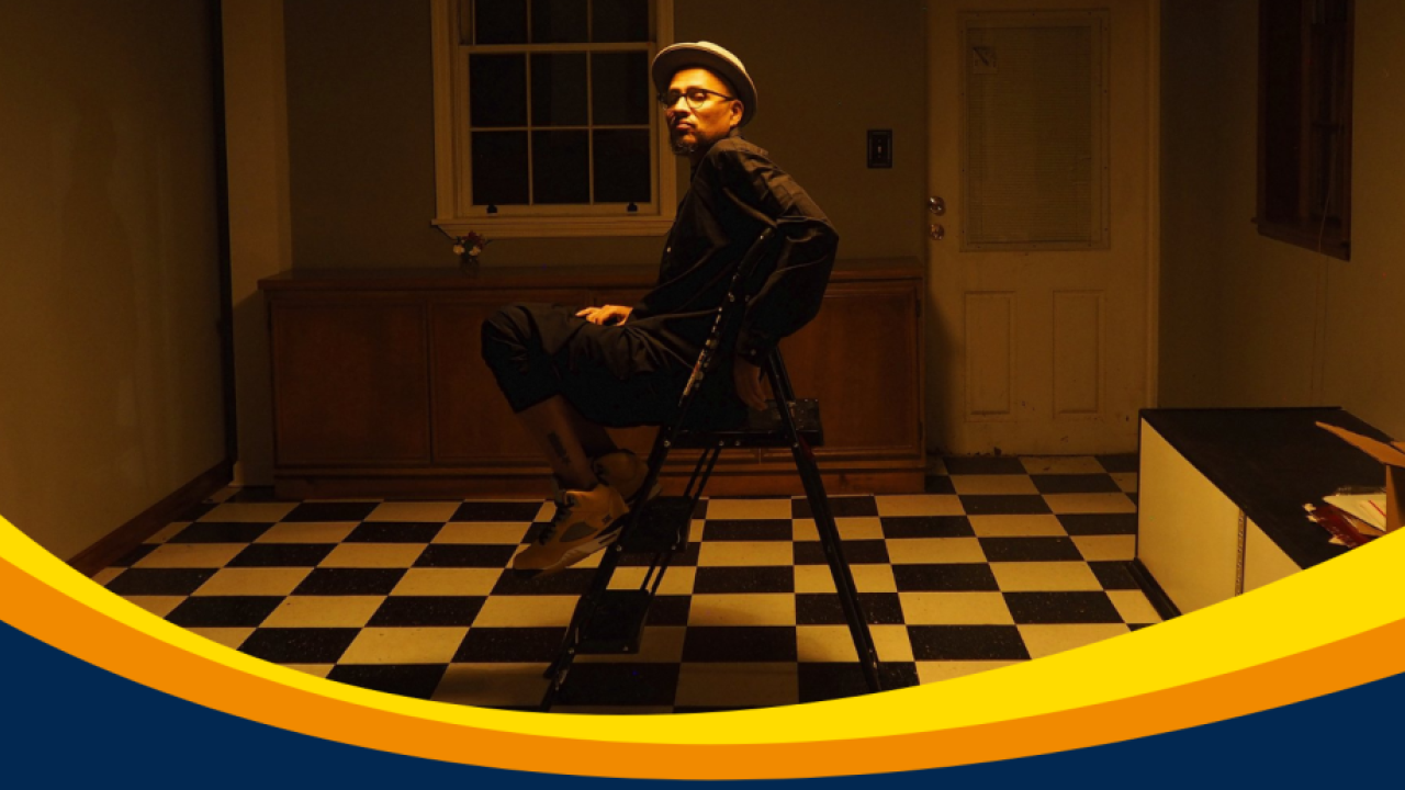 A picture of artist Bojafloca sitting in a chair looking towards the camera with dimly lit, yellow lighting in an empty house room