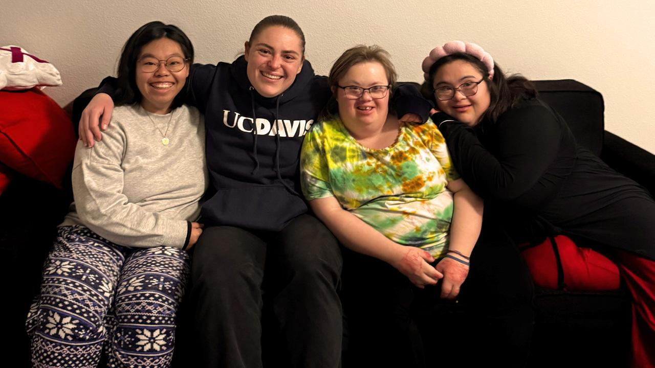 Four friends sitting closely on a couch, smiling; one in tie-dye shirt, another in sweatshirt with text.