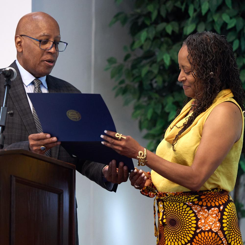 A man in a suit presents a certificate to a woman in a yellow top and patterned skirt at an indoor event.