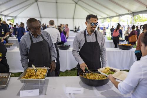 Two masked servers in aprons serving yellow rice to a guest under a white event tent