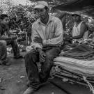 A farm laborer sits on a cot in the foreground, looking down. Two other workers sit in the shade of a tarp in the background.