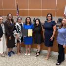 Group photo of eight smiling people in a wood-paneled chamber with US flag; one holds certificate