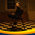 A picture of artist Bojafloca sitting in a chair looking towards the camera with dimly lit, yellow lighting in an empty house room