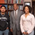 Three individuals pose together in an office setting, smiling. Bookshelves are in the background.