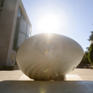 Close-up of the UC Davis Egghead sculpture with the sun shining brightly behind it, next to a campus building and surrounded by trees and benches.