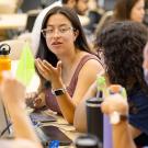 Students sit together at a table in a classroom, engaged in discussion with laptops and notebooks open, while one student gestures as she speaks.