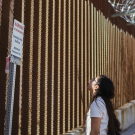 A woman gazes up at a tall border wall, sunlight illuminating her face.