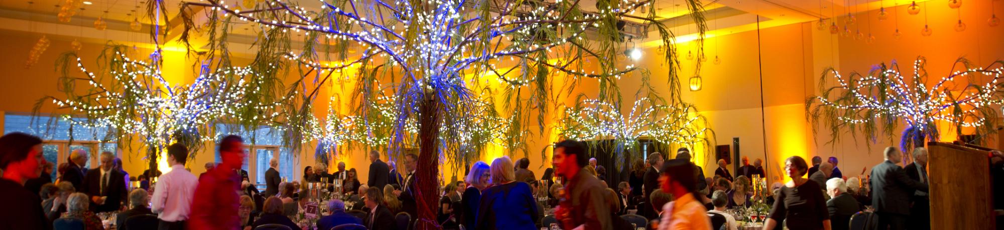 An indoor event space with decorative trees and a warm orange glow, filled with people at round tables.
