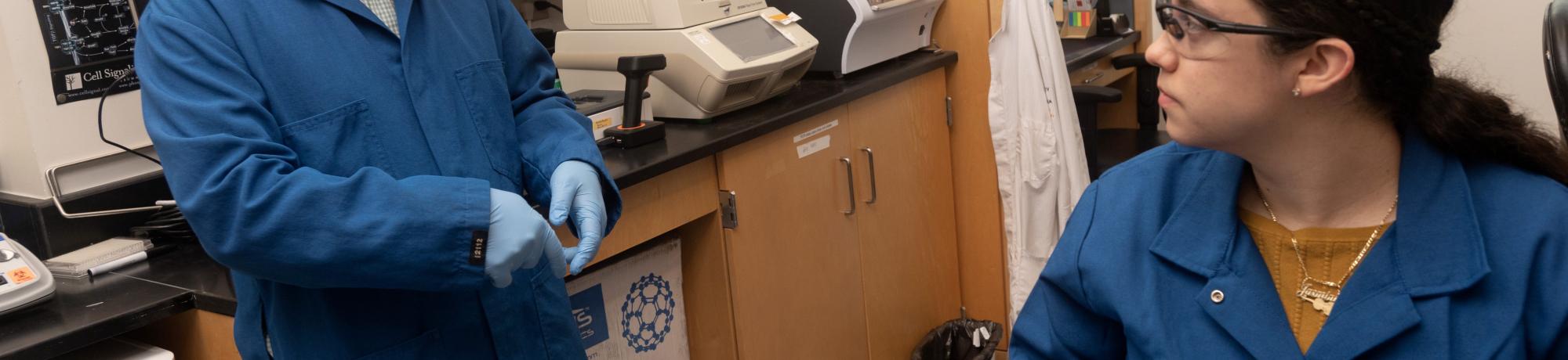 Luis Carvajal-Carmona, Ph.D., associate vice chancellor of academic diversity within the Division of Diversity, Equity and Inclusion, stands in his lab talking to one of his student lab assistants who is seated. Both are wearing a lab coat and lab goggles.